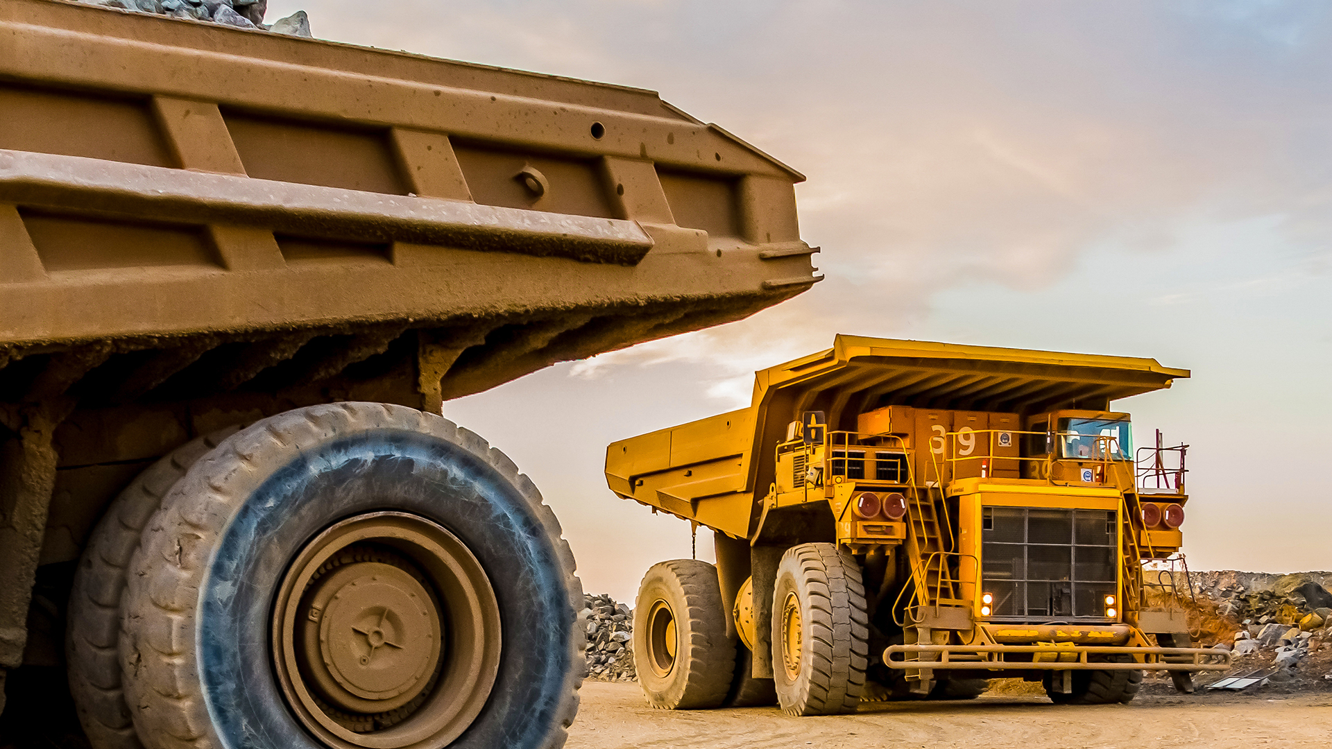 Two enormous yellow mining dump trucks in a quarry, with dusty ground and rock piles under a sunset sky.