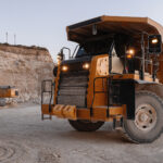 Large orange off-road dump truck at a rocky quarry, with an excavator and piles of dirt in the background.