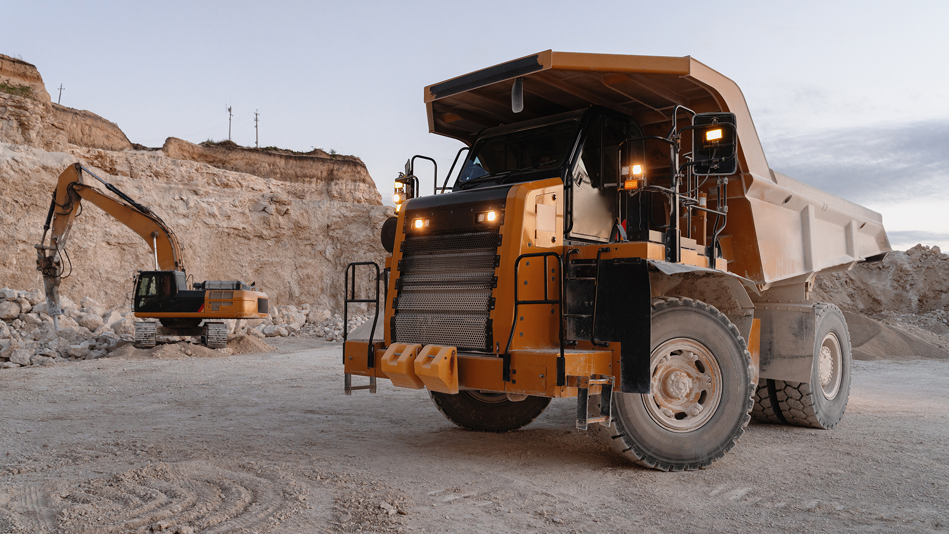 Large orange off-road dump truck at a rocky quarry, with an excavator and piles of dirt in the background.